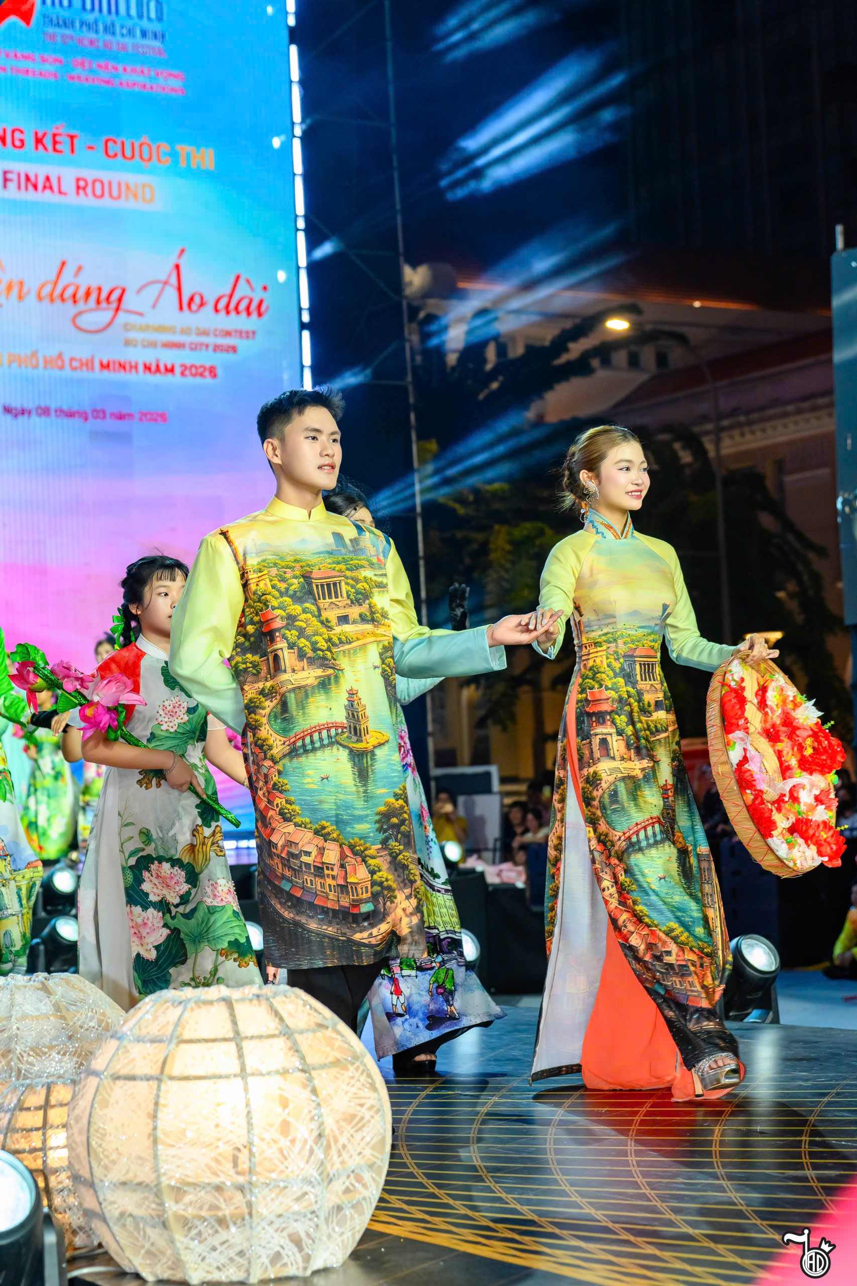 Một cặp thí sinh trong áo dài in cảnh Hồ Gươm với cầu Thê Húc và Tháp Rùa, phía sau là màn hình đề “Chung kết – Duyên dáng Áo dài Thành phố Hồ Chí Minh năm 2026”